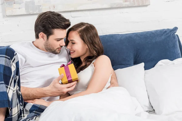 Hermosa pareja acostada en la cama, abrazando y sosteniendo regalo de cumpleaños en casa - foto de stock