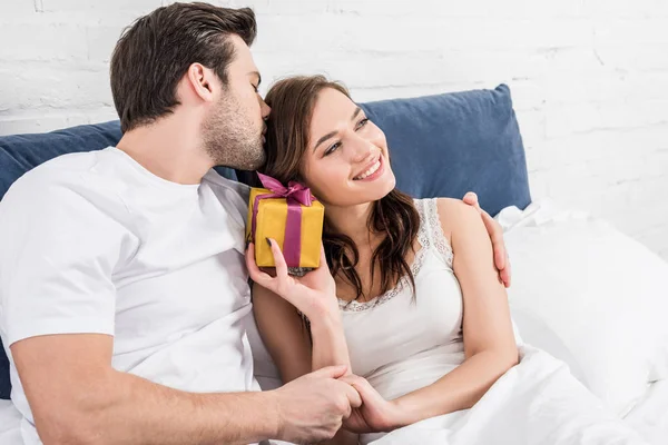 Pareja en ropa de dormir acostada en la cama, abrazando y sosteniendo regalo de cumpleaños en casa - foto de stock