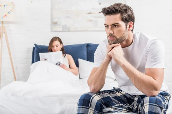 Dissatisfied man sitting on bed with folded hands while woman reading newspaper on background — Stock Photo