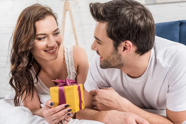 Feliz pareja acostada en la cama, sosteniendo regalo de cumpleaños y mirándose en casa - foto de stock