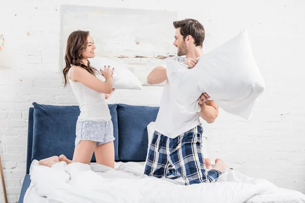 Casal feliz em pijama lutando com travesseiros no quarto — Fotografia de Stock