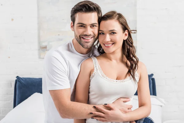 Schönes lächelndes Paar, das sich im Schlafzimmer umarmt und in die Kamera blickt — Stockfoto