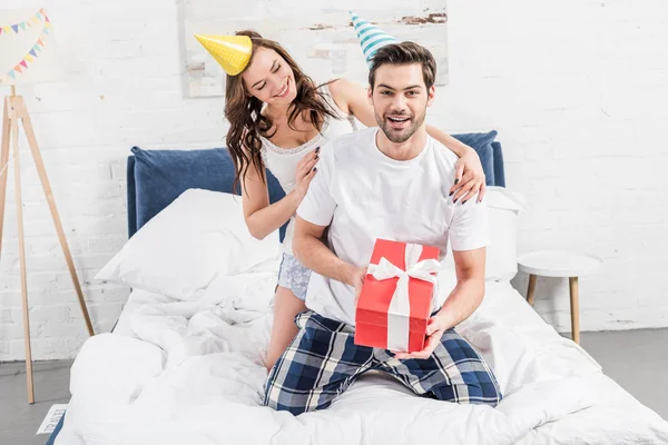 Счастливая пара в шляпах для вечеринок, празднующих событие и держащих подарок на день рождения в постели — стоковое фото