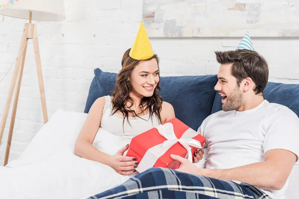 Couple in party lying hats in bed, holding birthday present and looking at each other — Stock Photo