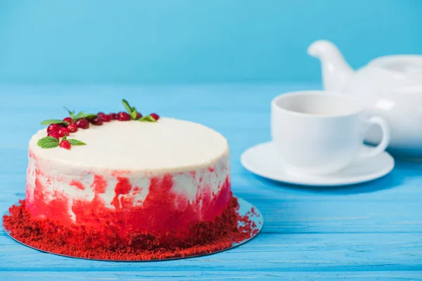 Gâteau décoré de groseilles rouges et feuilles de menthe près de tasse blanche et théière isolée sur bleu — Photo de stock