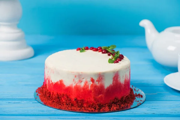 Торт, прикрашений червоною смородиною і листя м'яти між білою чашкою, чайним горщиком і стоїть ізольовано на синьому — стокове фото