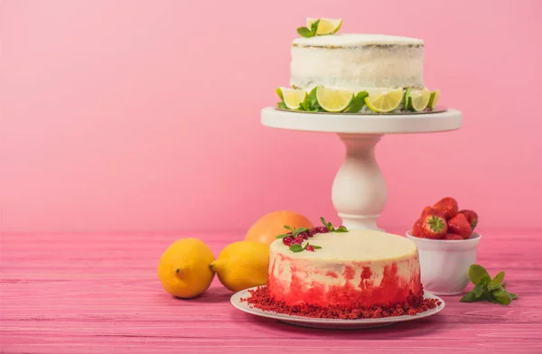 Pastel decorado con grosellas y hojas de menta cerca de frutas y pastel blanco con rodajas de limón en la superficie de madera rosa aislado en rosa - foto de stock