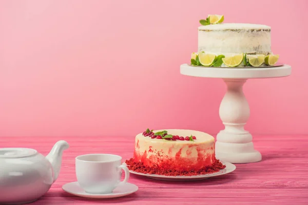 Kuchen dekoriert mit Johannisbeeren, Minzblättern und Limettenscheiben in der Nähe von Tasse und Teekanne auf Holzoberfläche isoliert auf rosa — Stockfoto