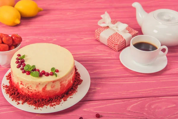 Close up of cake decorated with currants and mint leaves near tea pot, cup of strawberries and gift box on pink surface — Stock Photo