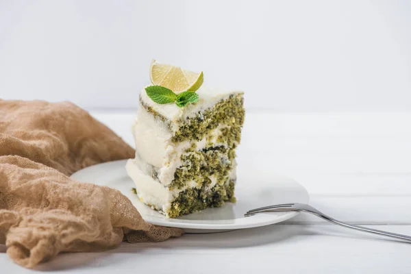 Pezzo di torta bianca decorato con fetta di lime e menta lasciare con forchetta su piattino isolato su bianco — Foto stock
