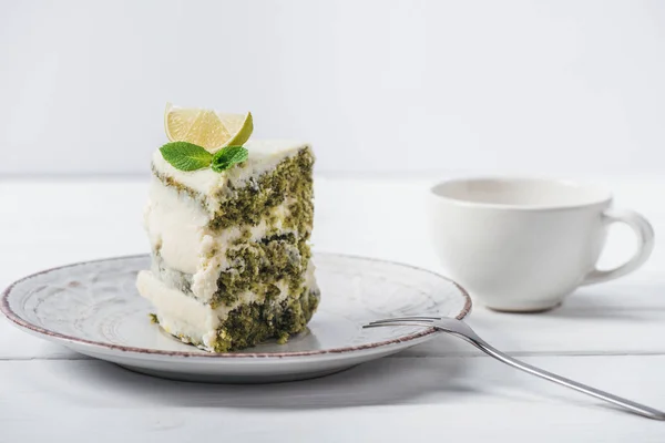 Pezzo di torta bianca decorata con fetta di lime e menta lasciare sul piattino vicino tazza isolata su bianco — Foto stock