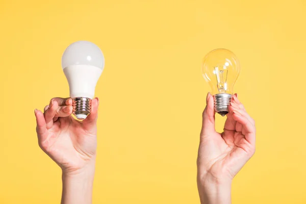 Vista recortada de manos femeninas eligiendo entre lámparas led y fluorescentes aisladas en amarillo, concepto de eficiencia energética - foto de stock