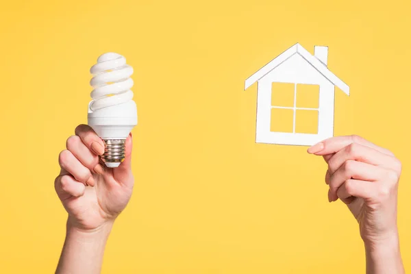 Обрезанный вид женских рук, держащих бумажный дом и флуоресцентную лампу в руках, изолированных на желтый, энергоэффективность в домашних условиях — стоковое фото