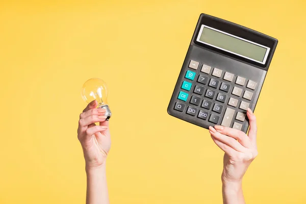 Vista recortada de la mujer sosteniendo lámpara led y calculadora en manos aisladas en amarillo, concepto de eficiencia energética - foto de stock