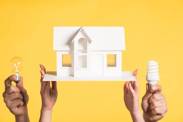 Обрізаний вид жінки, що тримає модель картонного будинку поблизу чоловіка з світлодіодними та люмінесцентними лампами, ізольовані на жовтому, енергоефективність в домашніх умовах концепція — стокове фото