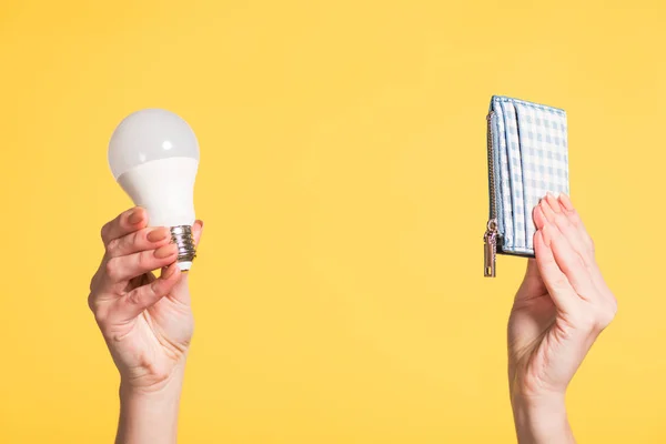 Vista recortada de mujer sosteniendo lámpara fluorescente y billetera en manos aisladas en amarillo, concepto de eficiencia energética - foto de stock