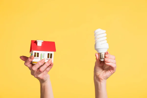 Vista recortada de la mujer sosteniendo lámpara fluorescente y modelo de casa en manos aisladas en amarillo, eficiencia energética en el concepto de casa - foto de stock
