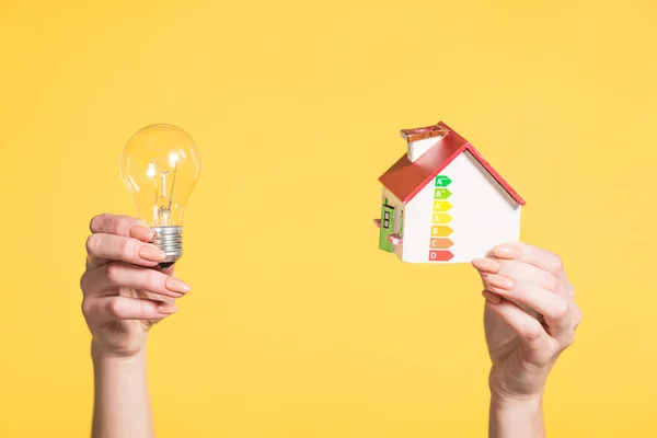 Vista recortada de la mujer sosteniendo lámpara led y modelo de casa en manos aisladas en amarillo, concepto de eficiencia energética en el hogar - foto de stock