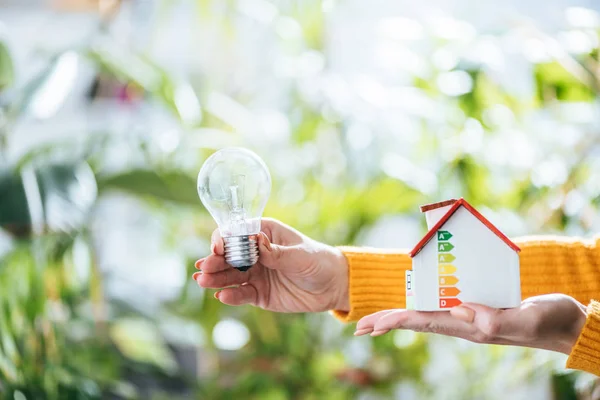 Селективная фокусировка свинцовой лампы и картонного дома модели в руках женщины, энергоэффективность в домашних условиях — стоковое фото