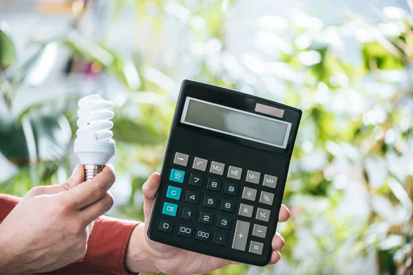 Vue recadrée de la lampe fluorescente et de la calculatrice entre les mains des hommes, concept d'efficacité énergétique — Photo de stock