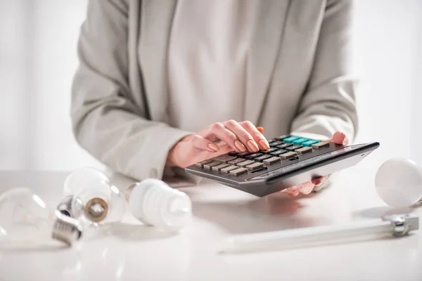 Обрезанный вид женщины с помощью калькулятора возле ламп на белом фоне, концепция энергоэффективности — стоковое фото