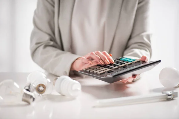 Vue recadrée de la femme à l'aide d'une calculatrice près des lampes sur fond blanc, concept d'efficacité énergétique — Photo de stock