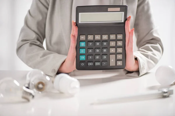 Vista ritagliata di donna in possesso di calcolatrice vicino a lampade su sfondo bianco, concetto di efficienza energetica — Foto stock