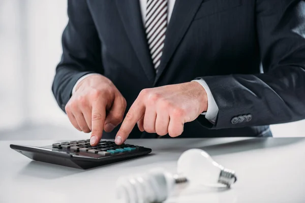 Vista recortada de hombre de negocios en traje usando calculadora cerca de lámparas fluorescentes sobre fondo blanco, concepto de eficiencia energética - foto de stock