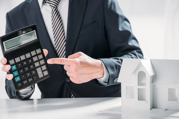 Abgeschnittene Ansicht des Geschäftsmannes, der mit dem Finger auf Taschenrechner in der Nähe von Kartonhaus auf weißem Hintergrund zeigt, Energieeffizienzkonzept — Stockfoto