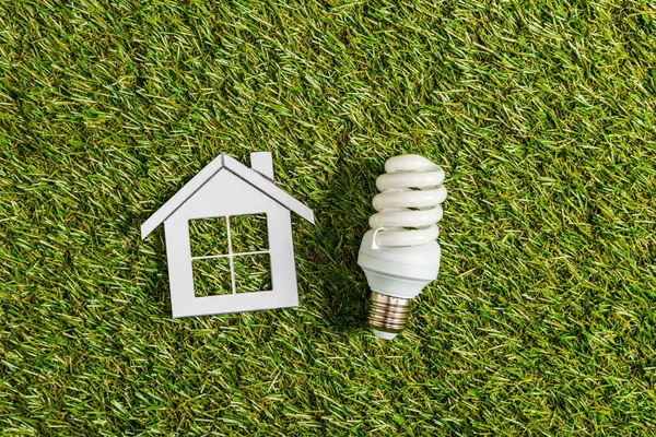 Вид зверху люмінесцентної лампи біля паперового будинку на зеленій траві, енергоефективність в домашніх умовах концепція — стокове фото