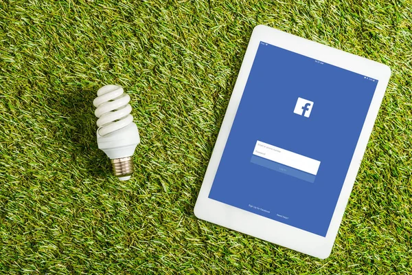 Vista superior da lâmpada fluorescente perto do tablet digital com aplicativo facebook na tela na grama verde, conceito de eficiência energética — Fotografia de Stock