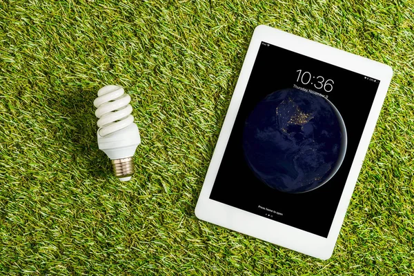Vista superior da lâmpada fluorescente perto do tablet digital com tela de bloqueio na grama verde, conceito de eficiência energética — Fotografia de Stock