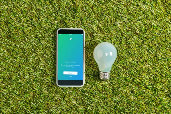 Top view of fluorescent lamp near smartphone with twitter app on screen on green grass, energy efficiency concept — Stock Photo