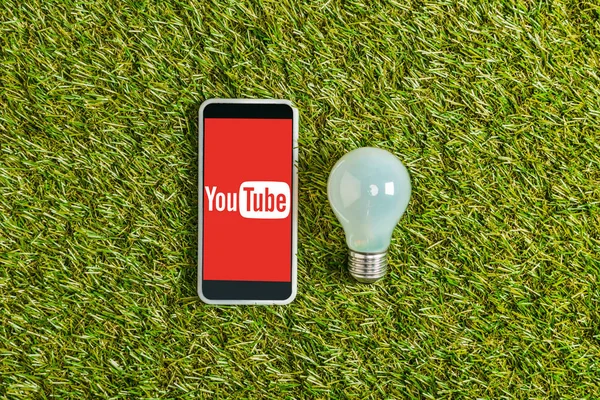 Vista superior da lâmpada fluorescente perto do smartphone com o aplicativo youtube na tela na grama verde, conceito de eficiência energética — Fotografia de Stock