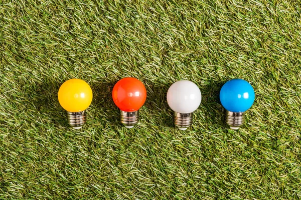 Colocação plana de lâmpadas fluorescentes coloridas na grama verde, conceito de eficiência energética — Fotografia de Stock
