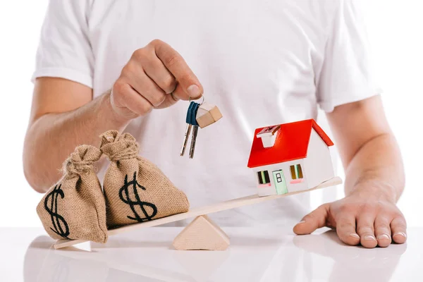 Vista parcial del hombre sosteniendo llaves cerca de moneybags y modelo de casa en escalas aisladas en blanco, concepto de hipoteca - foto de stock