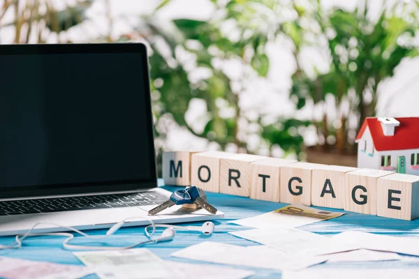 Enfoque selectivo de la computadora portátil con pantalla en blanco y llaves cerca de auriculares, billetes en euros y cubos de madera con letras en el escritorio, concepto de hipoteca — Stock Photo