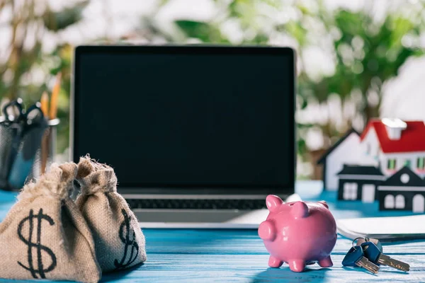Foco seletivo de sacos de dinheiro, banco piggy rosa e chaves na mesa de madeira com laptop no fundo, conceito de hipoteca — Fotografia de Stock