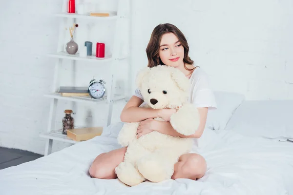 Belle femme étreignant ours en peluche et assis sur le lit avec literie blanche — Photo de stock