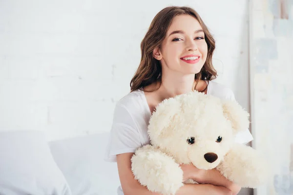 Selective focus of happy woman smiling and hugging teddy bear in bed — Stock Photo