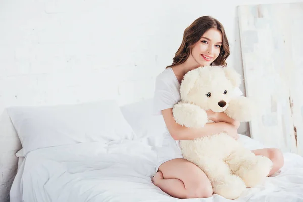 Beautiful woman hugging teddy bear and sitting on bed with white bedding — Stock Photo