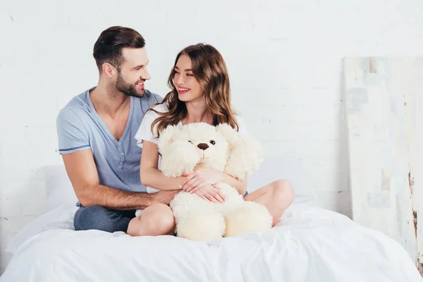 Jeune couple câlin ours en peluche tout en étant assis sur le lit avec literie blanche — Photo de stock