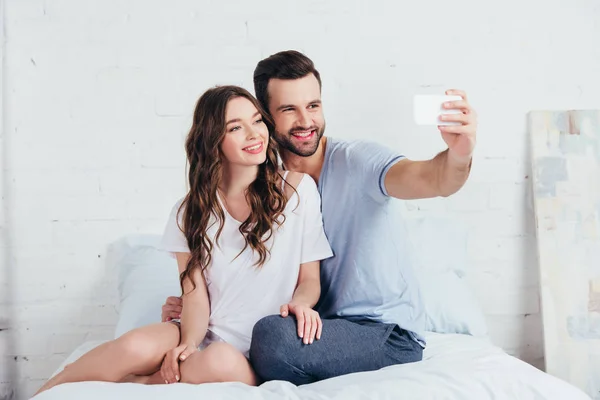 Подруга и парень сидят на кровати в спальне и делают селфи на смартфоне — стоковое фото