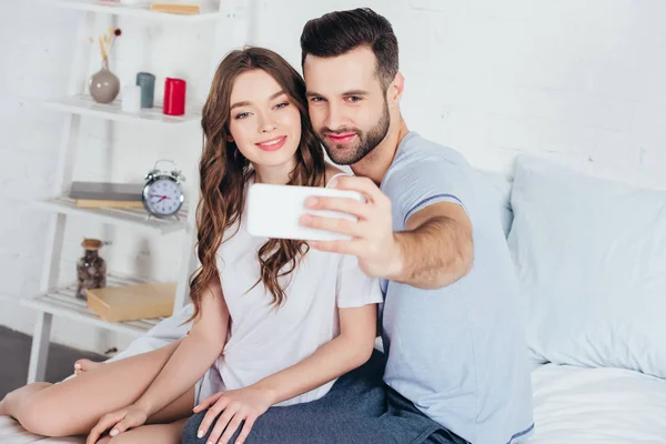 Молодая пара сидит на кровати в спальне и делает селфи на смартфоне — стоковое фото