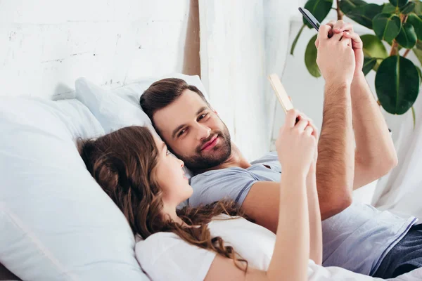 Pareja joven que se relaja en la cama mientras usa teléfonos inteligentes y se mira entre sí - foto de stock