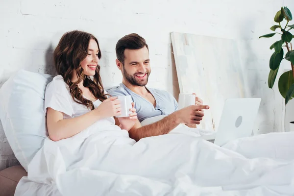 Молода пара тримає чашки, лежачи на м'якій білій постільній білизні і дивитися фільм на ноутбуці — стокове фото