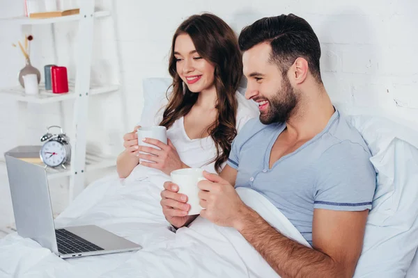 Young couple holding cups, lying in bed and watching film on laptop — Stock Photo