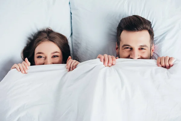 Novio y novia sonriendo y escondiéndose debajo de la manta en la cama - foto de stock