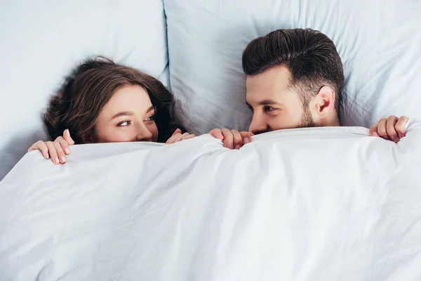 Joven pareja amorosa escondido bajo manta en la cama y mirando a los ojos - foto de stock