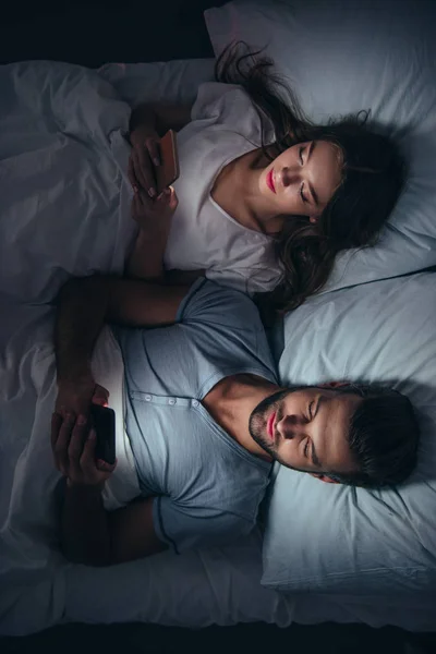 Jeune couple couché au lit et utilisant des smartphones la nuit — Photo de stock